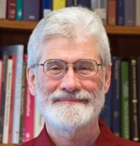 Formal headshot of faculty member Gerald Smith. Gerald is wearing glasses and a bookshelf with books is in the background.