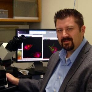 Headshot of faculty member Alex Paredez in lab space, in front of microscope and computer screen.