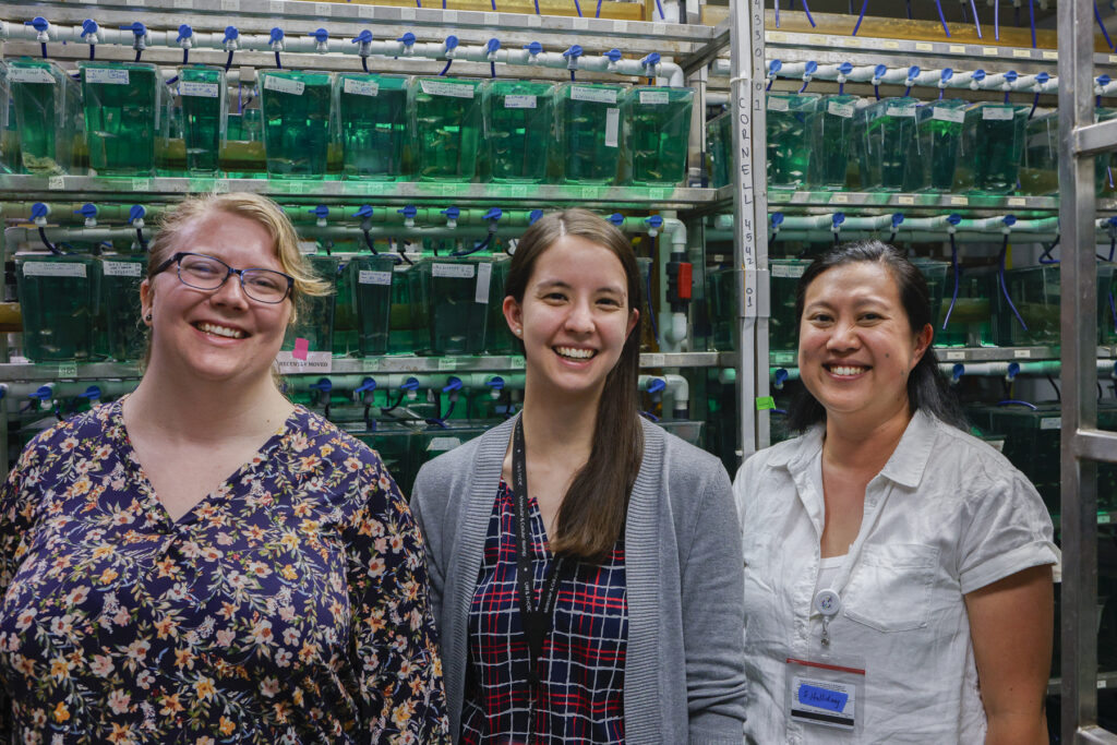 MCB student in front of rack of fish tanks in lab space. Two other lab mates stand on either side of her.