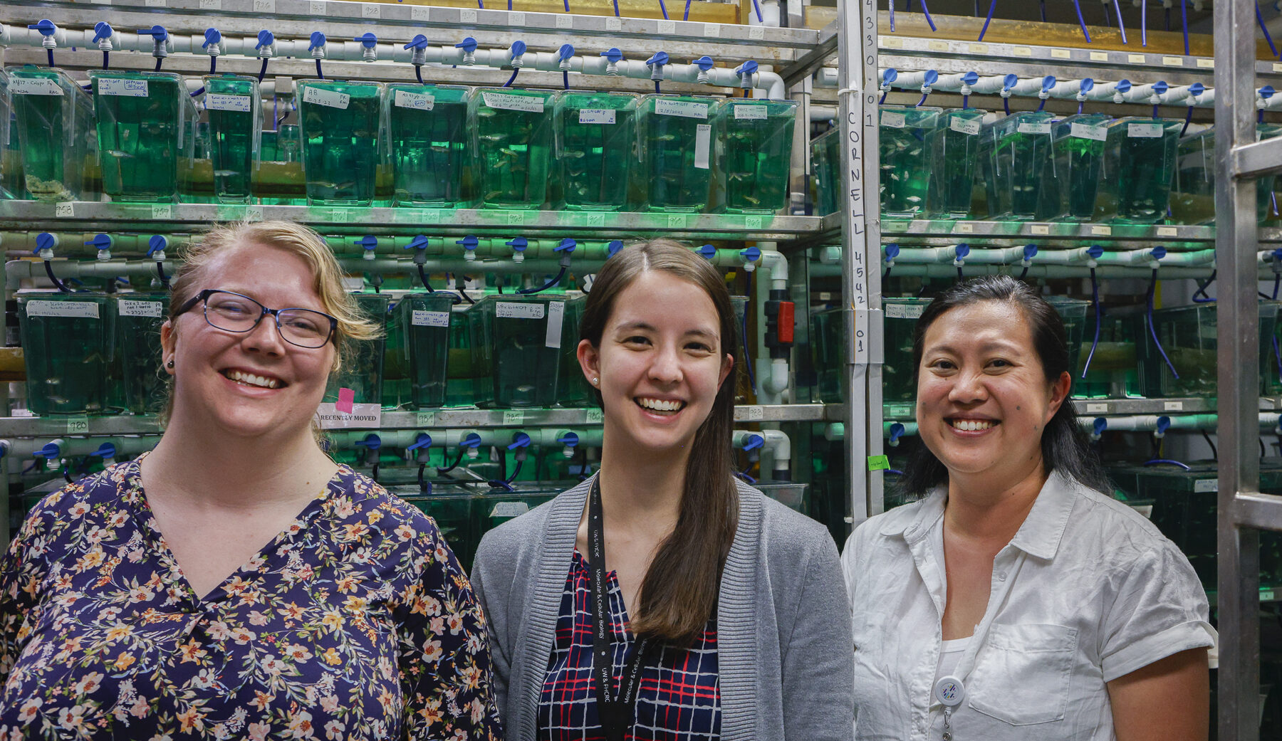 MCB student in front of rack of fish tanks in lab space. Two other lab mates stand on either side of her.