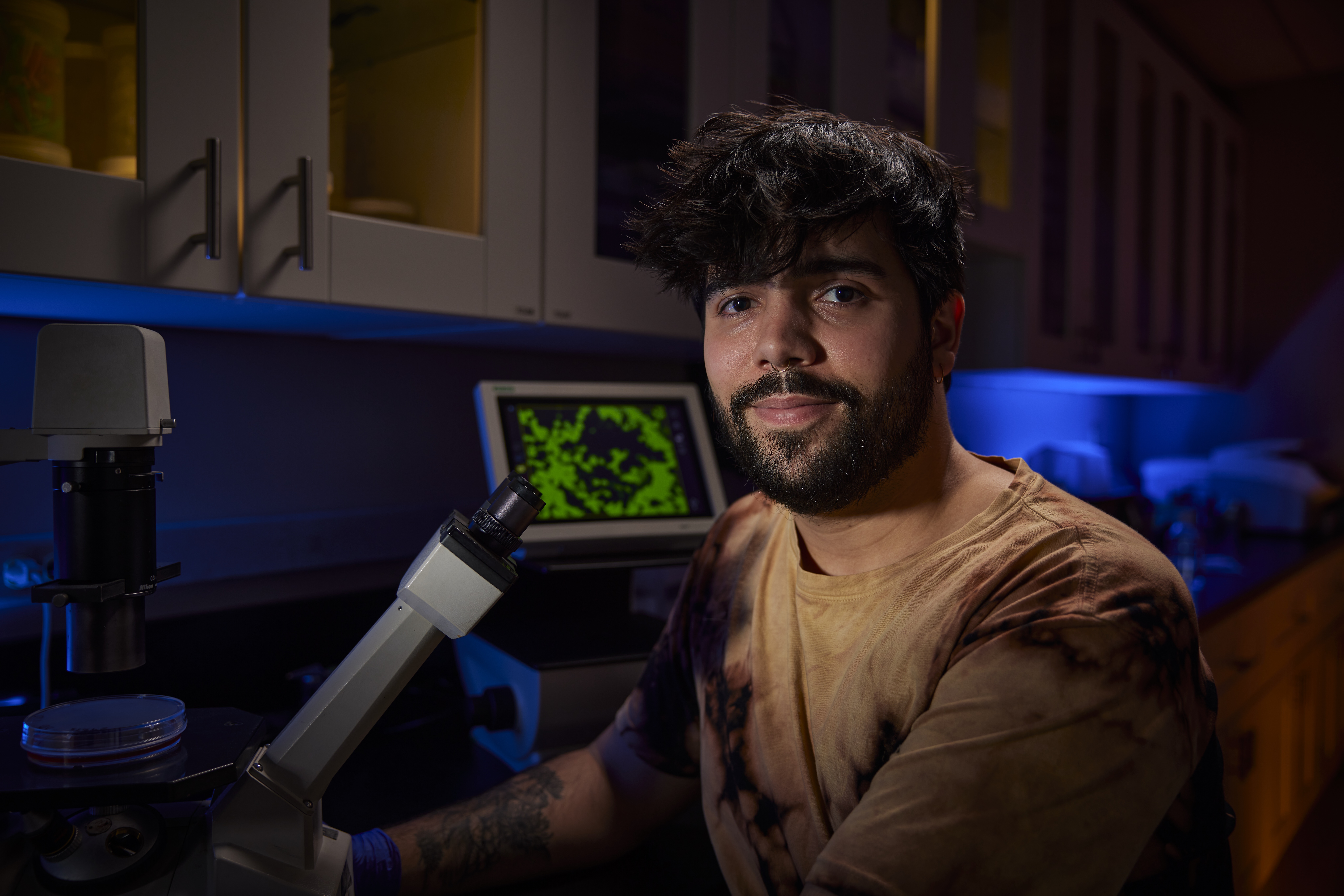 MCB student seated at lab desk next to microscope and looking at camera.