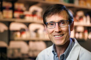 Formal headshot of faculty member William Grady. Dr. Grady is smiling in glasses and a white lab coat standing in a blurred laboratory.